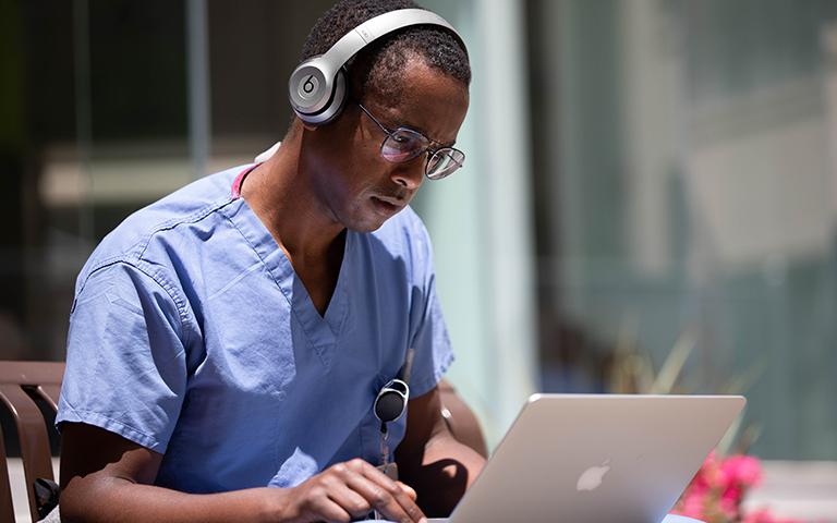 Man in medical scrubs sitting outside at daytime at their laptop